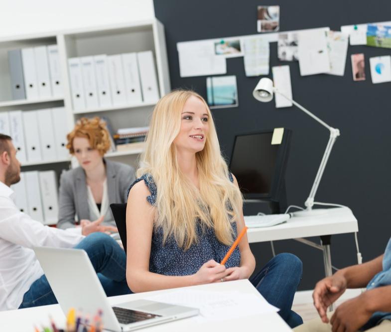 Two young businesspeople in a busy office sitting chatting with a beautiful young blond woman and young African American male in the foreground and colleagues working behind-2